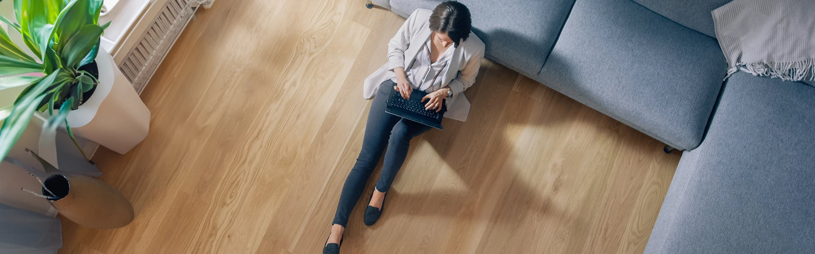 Eine Frau sitzt auf ihrem neuen Vinylboden angelehnt an ein blaues Sofa und hat einen Laptop auf dem Schoß