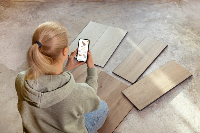 Eine blonde Frau sitzt auf dem Boden, vor sich mehrere Fußbodenmuster zum Testen und Prüfen ausgelegt, in den Händen hält sie ein Smartphone auf dem weitere Vinylböden abgebildet sind