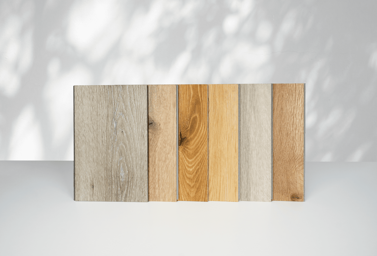 Im Vordergrund stehen sechs unterschiedliche Musterdielen unseres Vinyls aufgereiht. Im Hintegrund sieht man eine weiße Wand auf der sich Schatten von Pflanzen abbilden.