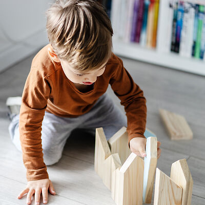 Ein kleiner Junge mit orangenem Oberteil und grauer Hose spielt konzentriert mit Holzklötzen auf einem lichtdurchflutetem hellgrauen Vinylboden