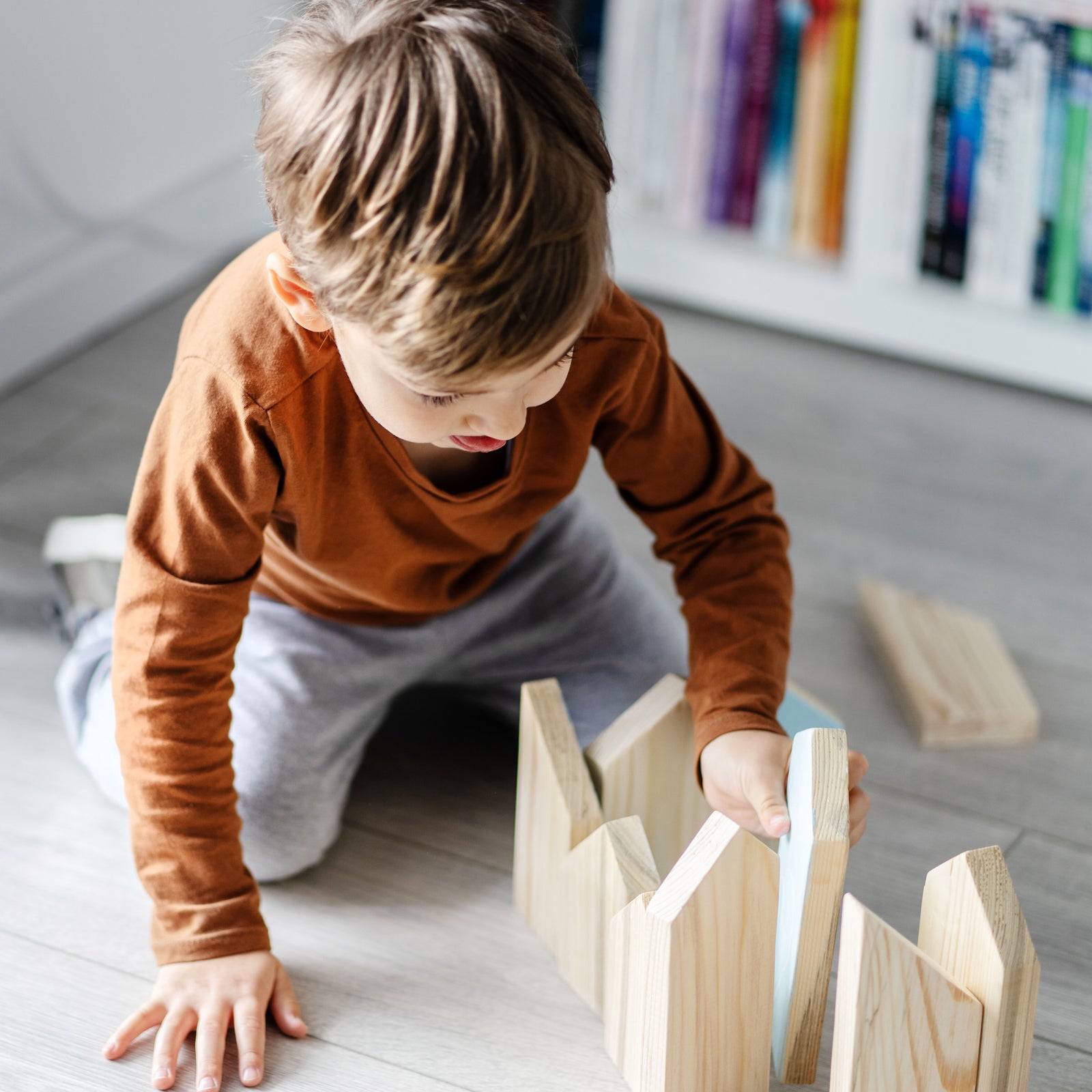 Ein kleiner Junge mit orangenem Oberteil und grauer Hose spielt konzentriert mit Holzklötzen auf einem lichtdurchflutetem hellgrauen Vinylboden