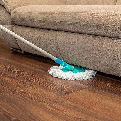 Auf einem dunkelbraunen Vinylboden wischt ein Wischmopp unter einem hellgrauen Sofa in einer Wohnung